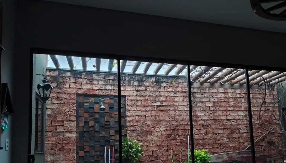 Vista de dentro de uma sala através da porta de vidro com película: a parede de tijolos e o pergolado da área externa aparecem nítidos.