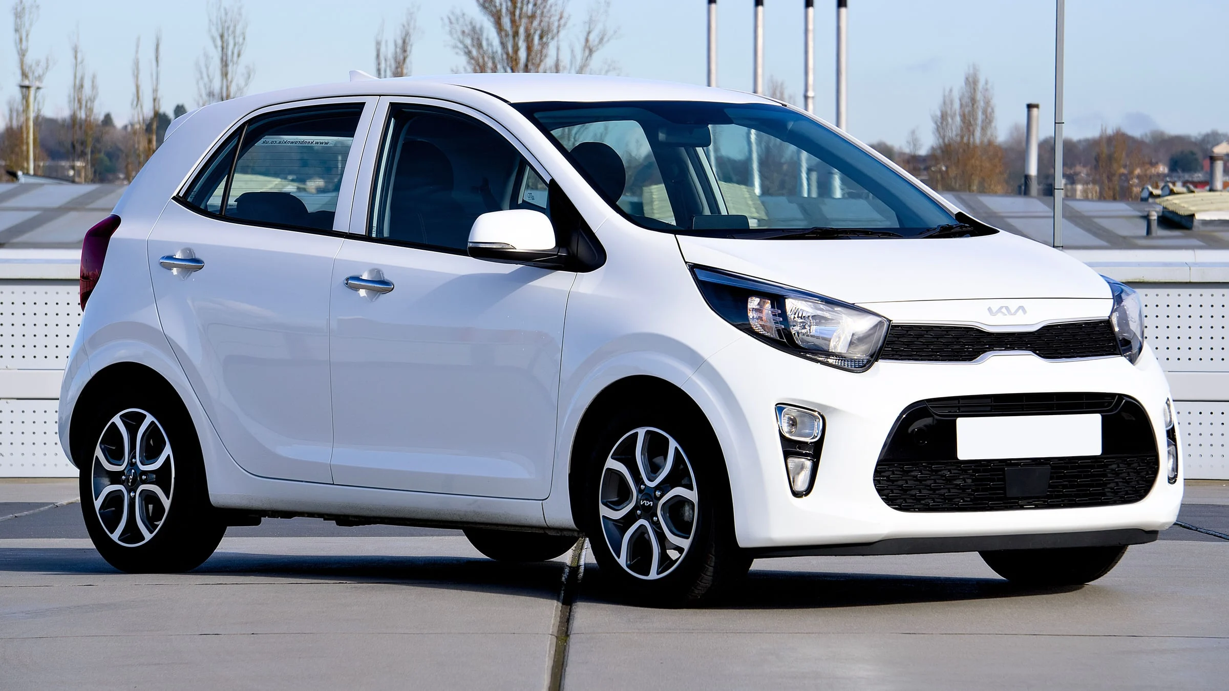 Kia Picanto branco, hatch de cinco portas, visto em três quartos de frente pelo lado direito, estacionado numa cobertura em dia de sol, com a película simulada nos vidros.