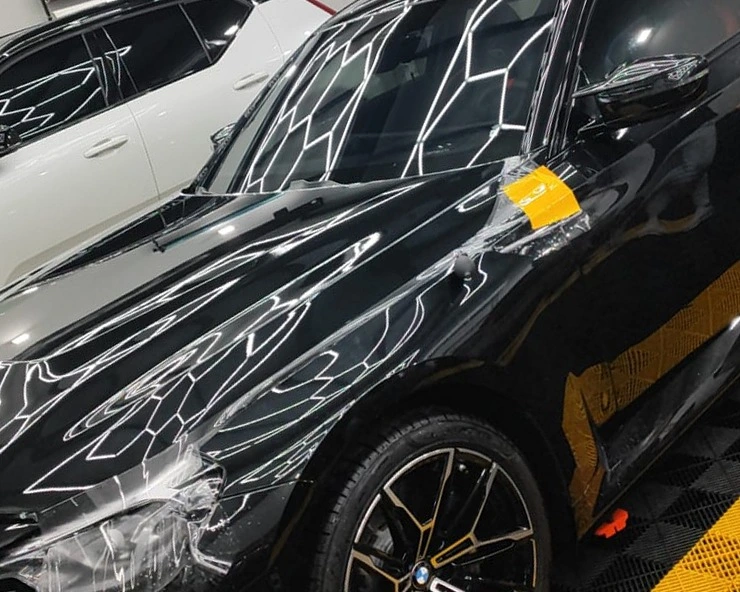 Película de proteção sendo aplicada sobre o capô e o para-lama de um carro preto, presa com fita, com o farol e a roda dianteira em quadro e um segundo carro ao fundo do box.