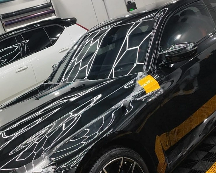 Película de proteção sendo aplicada sobre a lateral de um carro preto, presa com fita, com o restante da lataria ainda exposto e um segundo carro ao fundo do box.