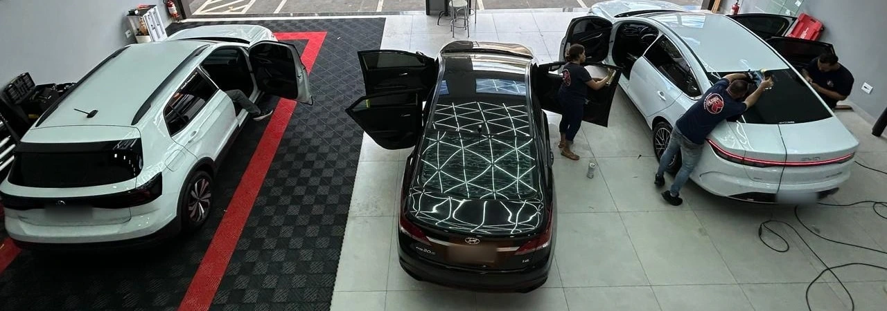 Vista de cima do salão da RFilmes: três carros com as portas abertas e três aplicadores trabalhando na película dos vidros, um em cada veículo.